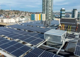 Rooftop solar panels cover a building as workers in safety gear install equipment with a city skyline in the background.