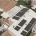 Aerial view of a white building roof covered with multiple solar panels, with workers in hard hats installing them and surrounding houses and cars visible in the neighborhood.